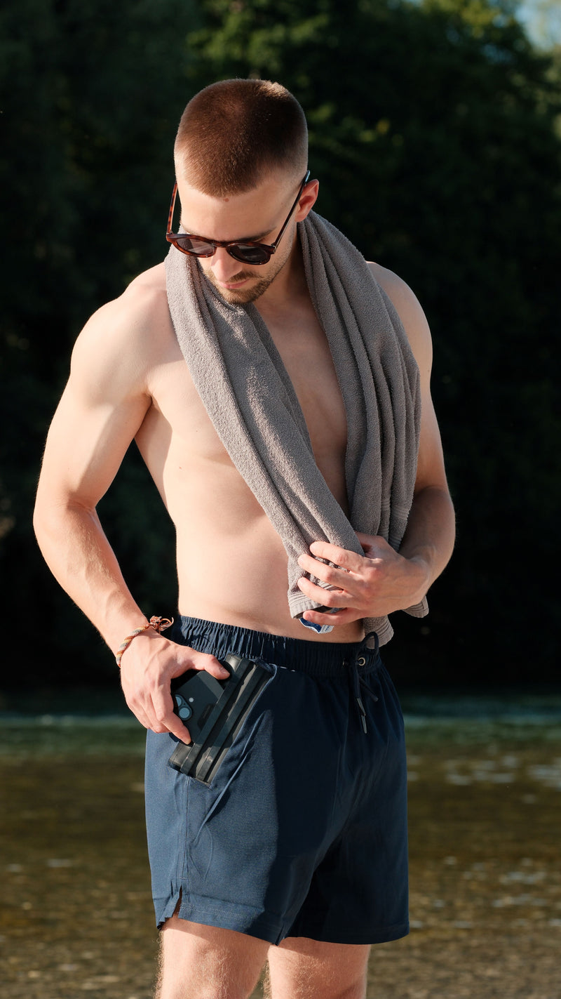 Model steckt ein Smartphone in die integrierte wasserdichte Tasche der BEVA Badehose am See, sicherer Schutz für Handy und Wertsachen beim Schwimmen.